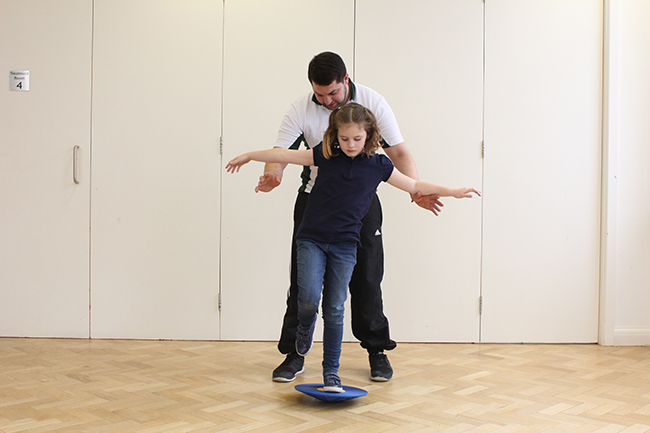 Child balancing on wobble board, therapist behind offering support