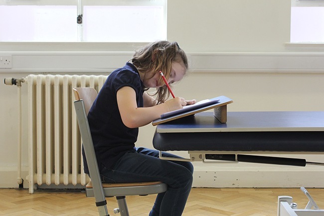 Child sitting on posturite chair, writing on a sloped writing board